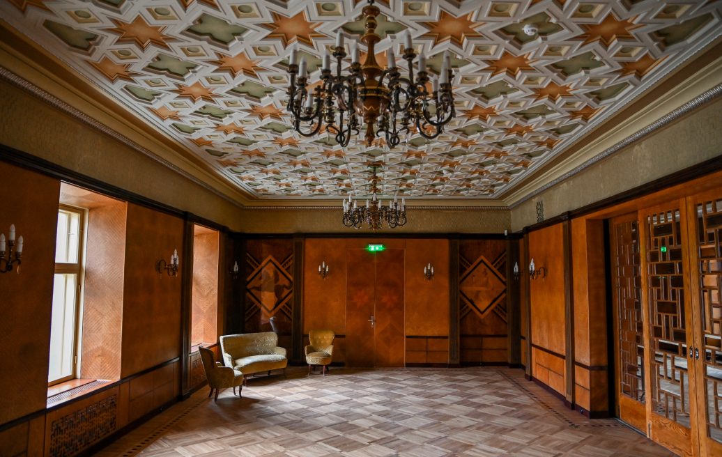 Siguldas Jaunās pils galvenā telpa ar divām lustrām un greznu griestu dizainu no zaļiem un oranžiem elementiem