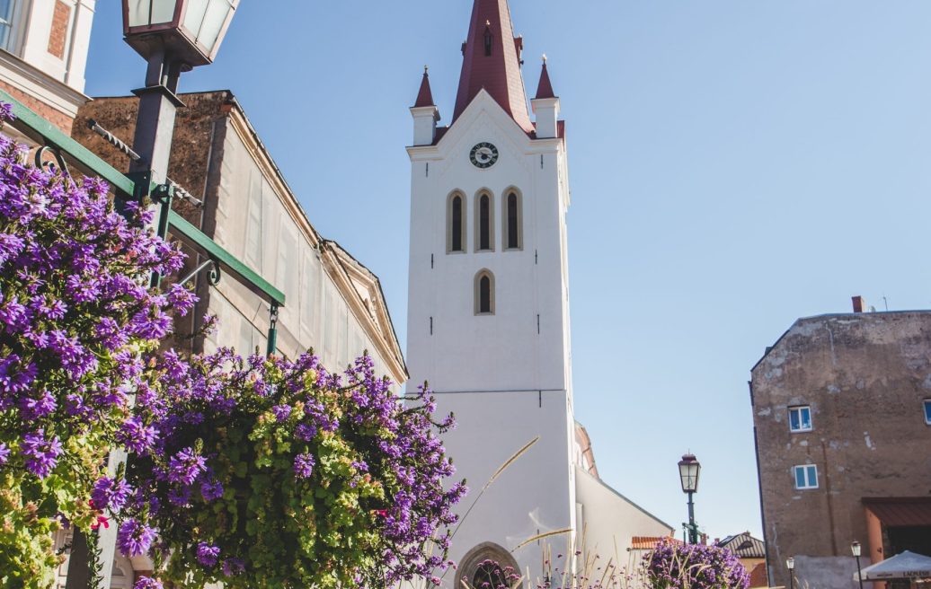 Cēsu Sv. Jāņa baznīcas ēka no ārpuses ar pilsētu fonā un violetiem ziediem pilsētas laukumā
