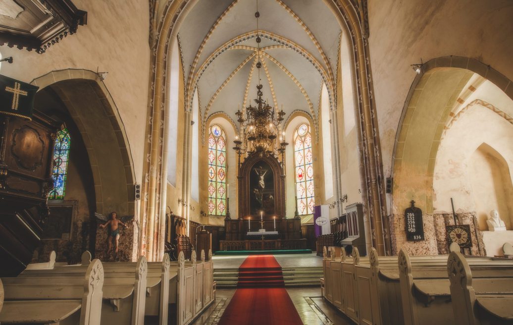 Cēsu Sv. Jāņa baznīcas altārs, kur iedegtas trīs svētās sveces, redzama lustra, sēdvietas sānos, sarkans paklājs uz zemes